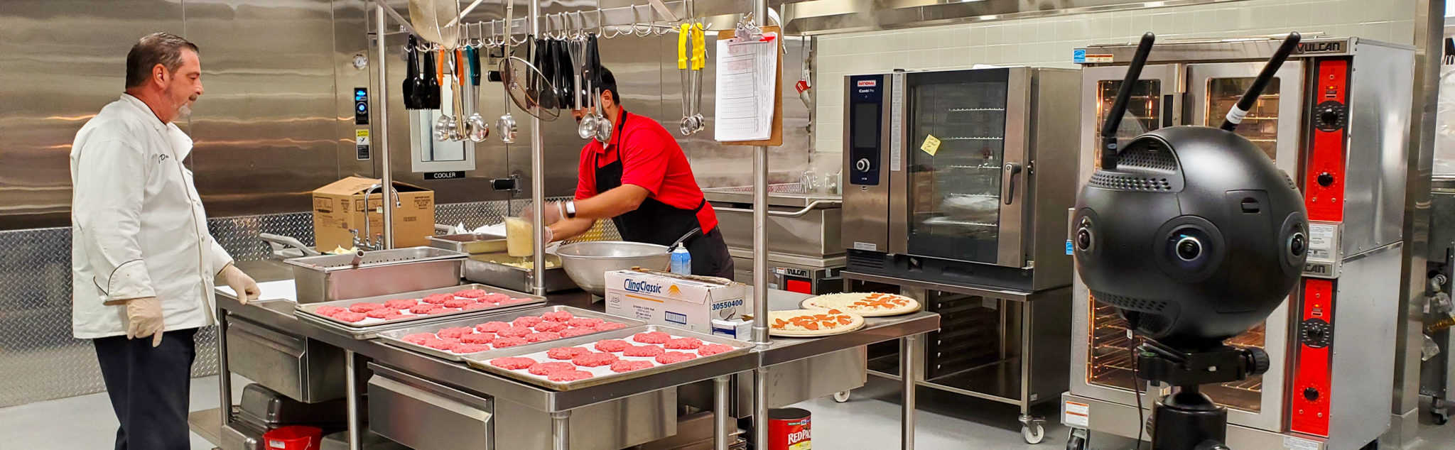 two chefs preparing fresh meals in a school kitchen with a 360 video camera